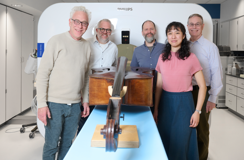 Members of the instrument imaging team stand on either side of a double bass that lays on a table in front of a CT machine.